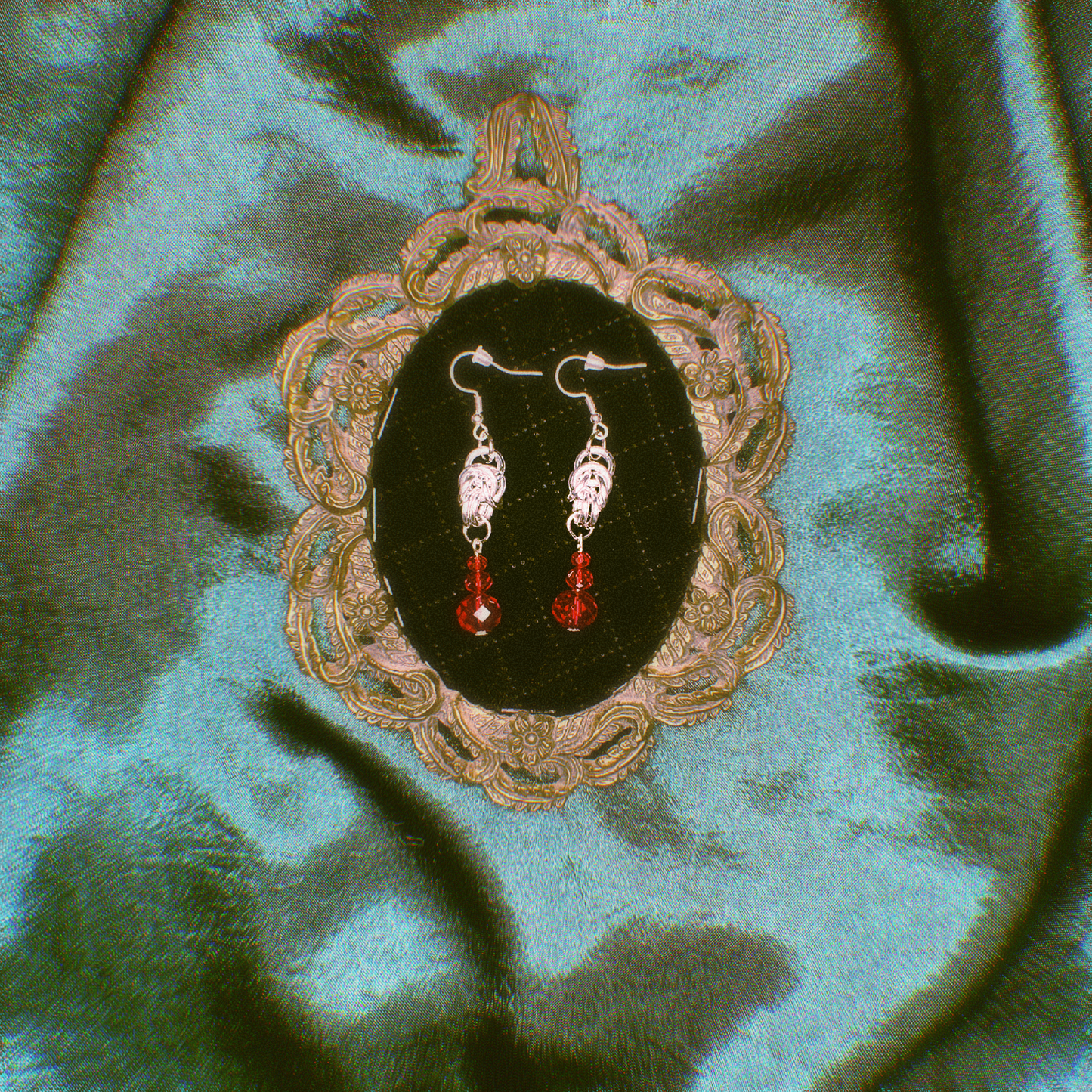 Pair of earrings with red stones displayed in an ornate frame on a green fabric background